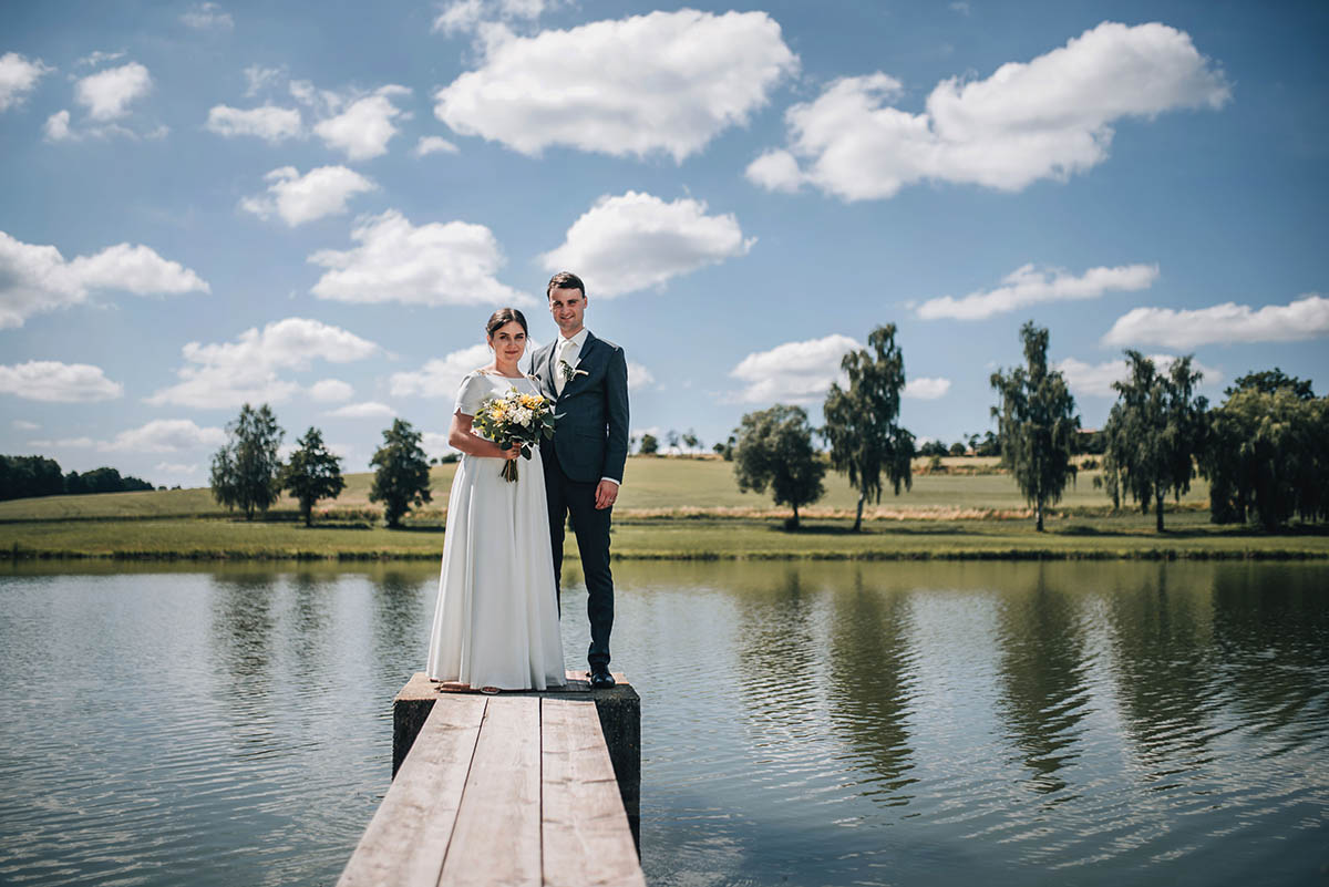 Novomanželé stojící na kraji mola u rybníka.