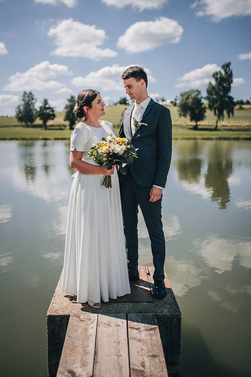 Novomanželé stojící na lávce u rybníka s bílými mraky v pozadí.